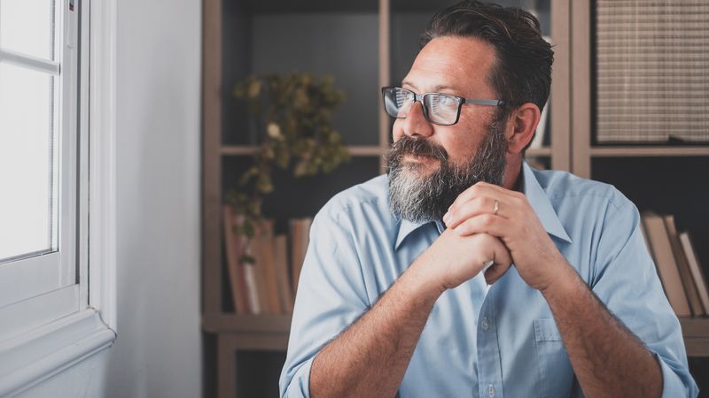 An adult with a beard and glasses is sitting in front of an open laptop in a home office and looking out of the window