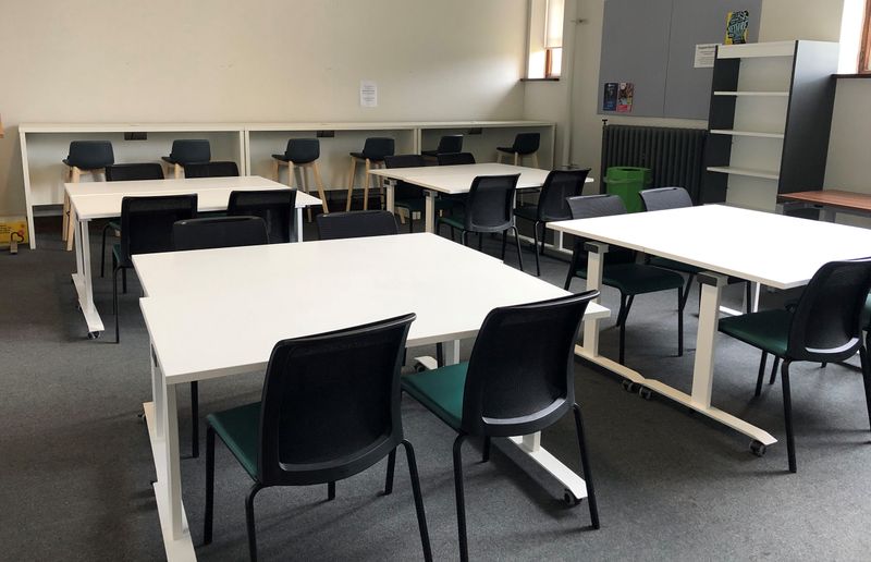 A room set up like a classroom filled with white desks and black chairs. Bookshelves line the walls 