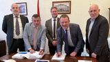 Five men in suits smile to the camera, two at the front are leaning over and signing a big document.