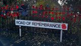 A photograph of a road sign which says 'road of remembrance'. It is infront of a fence and a green bush. There are poppies on the fence.