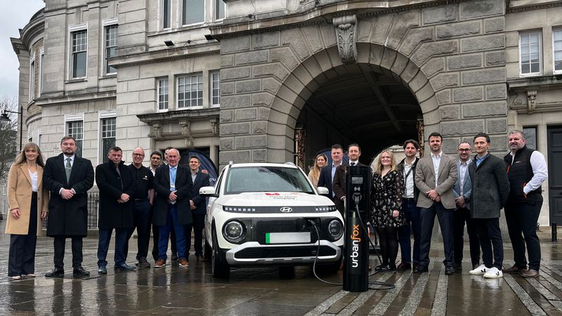 A group of people are standing infront of a large building. They are standing around an electric car and an electric charger which says 'Urban Fox'. 