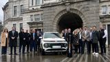 A group of people are standing infront of a large building. They are standing around an electric car and an electric charger which says 'Urban Fox'. 