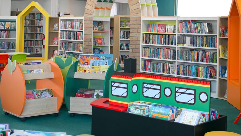 Bright and colourful children&rsquo;s library area with low book displays shaped like playful objects, a train‑themed bookshelf, and tall shelves of books in the background. A decorative archway stands at the centre, with a variety of children&rsquo;s picture books and chapter books arranged throughout the space.