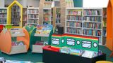 Bright and colourful children&rsquo;s library area with low book displays shaped like playful objects, a train‑themed bookshelf, and tall shelves of books in the background. A decorative archway stands at the centre, with a variety of children&rsquo;s picture books and chapter books arranged throughout the space.
