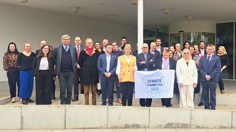 A large group of people standing in several rows outside a modern building entrance. Some individuals at the front hold a banner reading &ldquo;Straits Committee,&rdquo; which features a circular logo and wave motif. The group is dressed in formal and business-style clothing.