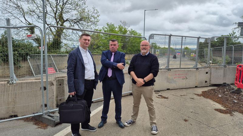 Three people standing on a pavement beside temporary metal fencing at a roadworks site, with concrete barriers, trees, and an overcast sky in the background