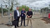 Three people standing on a pavement beside temporary metal fencing at a roadworks site, with concrete barriers, trees, and an overcast sky in the background