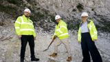Three people in high‑visibility jackets and safety helmets stand on a chalk hillside at a construction site, with one person holding a spade to mark the start of works.
