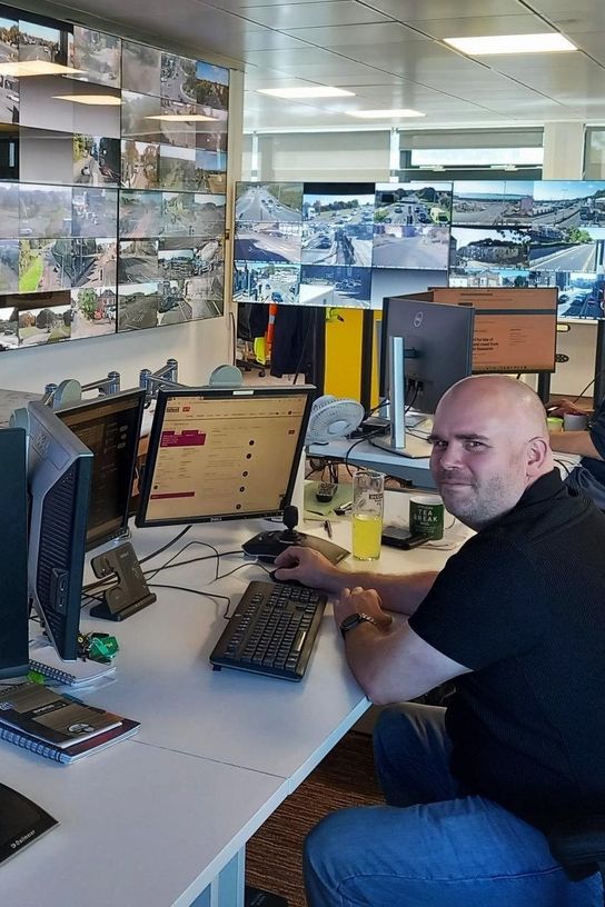 &ldquo;Person seated at a desk in a traffic control room, working at a computer workstation, with multiple wall-mounted screens displaying live road and junction camera feeds in the background.&rdquo;