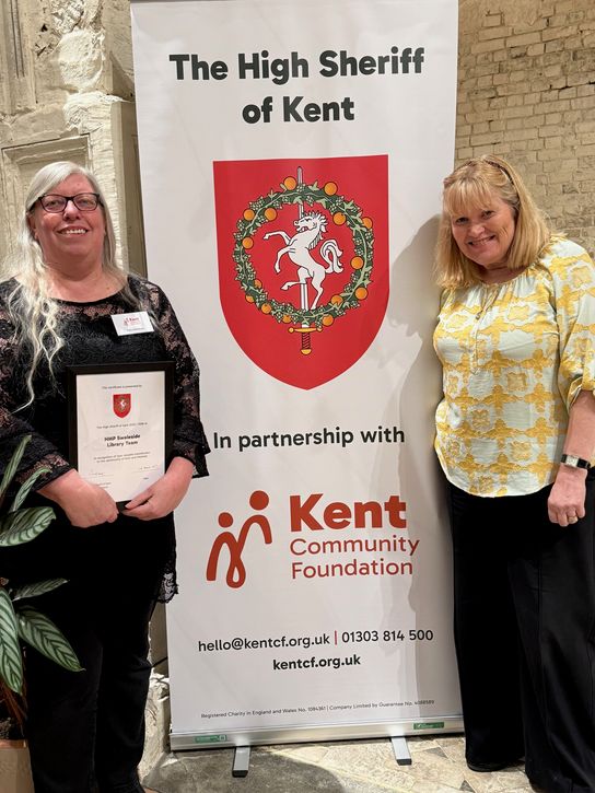 Two people pose on either side of a large banner reading &ldquo;The High Sheriff of Kent,&rdquo; featuring a red crest with a white horse. The person on the left holds a framed certificate for &ldquo;HMP Swaleside Library Team.&rdquo; The banner also includes &ldquo;Kent Community Foundation&rdquo; and contact details at the bottom.
