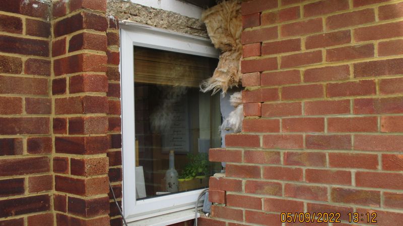 A brick wall with a partially dismantled section around a white-framed window, exposing insulation and construction materials 
