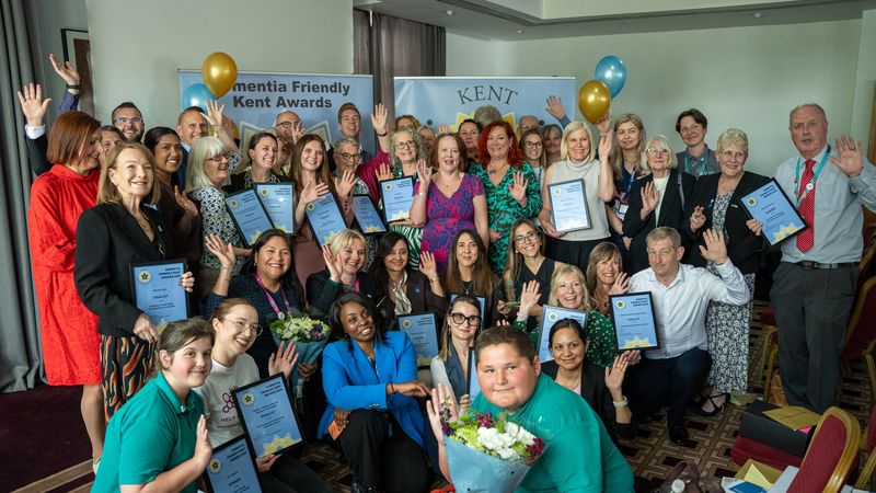 A group of people, some holding certificates and balloons, at the Dementia Friendly Kent Awards, gathered around banners and smiling for the camera