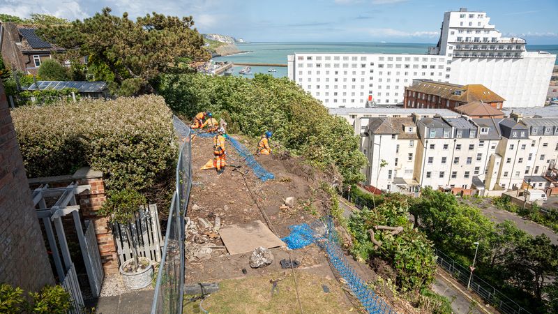 Workers in high-visibility orange clothing install protective netting along a steep cliffside above Folkestone, with buildings and the sea visible in the background.