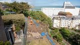Workers in high-visibility orange clothing install protective netting along a steep cliffside above Folkestone, with buildings and the sea visible in the background.