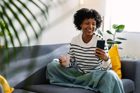A smiling woman sat on a sofa with a mobile phone in one hand and a mug in the other.