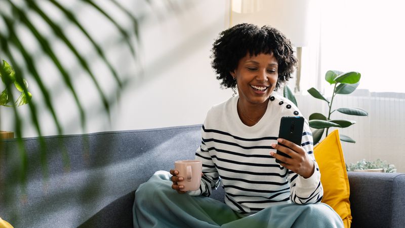 A smiling woman sat on a sofa with a mobile phone in one hand and a mug in the other.