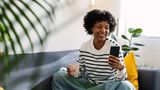 A smiling woman sat on a sofa with a mobile phone in one hand and a mug in the other.