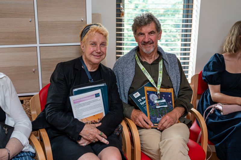 Two attendees seated indoors at a community event, holding newsletters and information packs