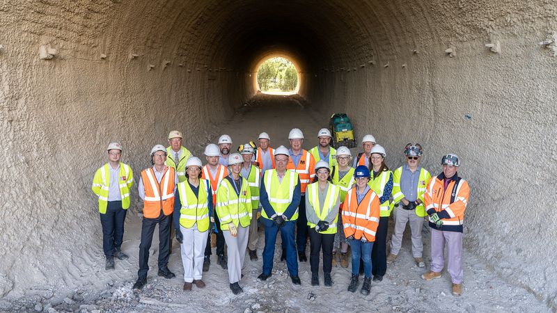 Group of construction and project staff wearing high-visibility clothing and hard hats standing inside a large underpass, with daylight visible at the far end.