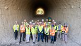 Group of construction and project staff wearing high-visibility clothing and hard hats standing inside a large underpass, with daylight visible at the far end.