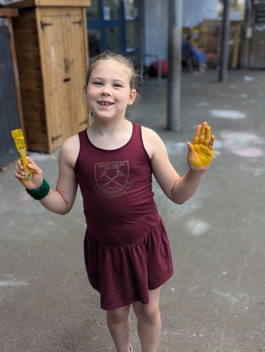 A child standing outdoors holds a paintbrush covered in yellow paint and raises a paint-covered hand towards the camera. The child is wearing a maroon sleeveless dress with a West Ham United logo on the front. The background shows a school or playground area with chalk markings on the ground and wooden structures nearby.