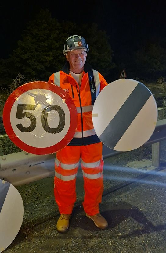 Peter Osborne in orange high-visibility clothing and a hard hat holding an old 50mph speed limit sign and a new national speed limit sign beside a roadside barrier at night.