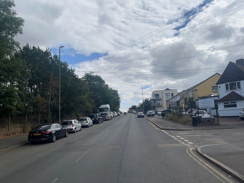 View looking along the A226 London Road in Stone, Dartford. A two-lane road rises gently uphill with parked cars lining the left-hand side. Trees and fencing run alongside the road to the left, while commercial buildings and a side road, Oak Road, are visible on the right. Overhead utility cables cross a cloudy sky.