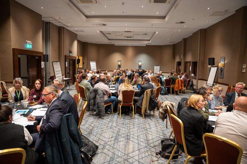 Delegates seated at round tables take part in group discussions during a large stakeholder workshop. The conference room is arranged for collaborative sessions, with participants working together around multiple tables.