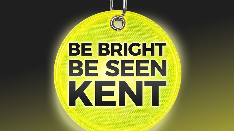 Bright fluorescent yellow reflective circular tag hanging from a metal key ring against a dark background. The tag displays the words &ldquo;BE BRIGHT BE SEEN KENT&rdquo; in large black capital letters and has a reflective pattern designed to improve visibility in low light conditions.