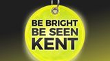 Bright fluorescent yellow reflective circular tag hanging from a metal key ring against a dark background. The tag displays the words &ldquo;BE BRIGHT BE SEEN KENT&rdquo; in large black capital letters and has a reflective pattern designed to improve visibility in low light conditions.