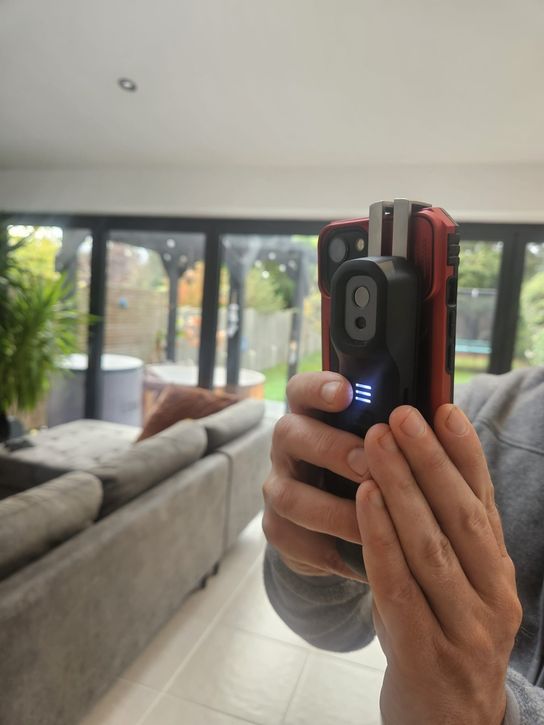 A thermal imaging camera attached to a smartphone, held in a living room with large glass doors overlooking a garden.
