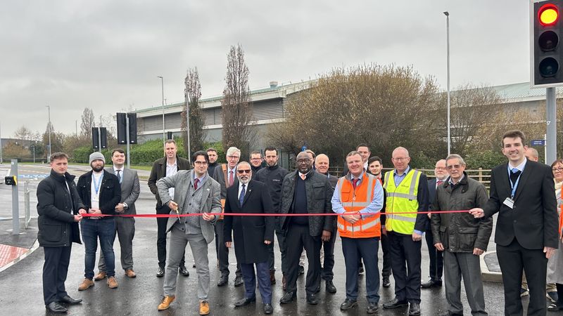 A group of people stand on a rainy road. They are holding a ribbon and one person in the centre who is about to cut the ribbon.