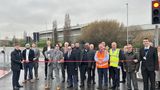 A group of people stand on a rainy road. They are holding a ribbon and one person in the centre who is about to cut the ribbon.