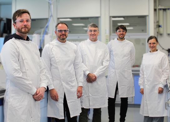 A group of five adult scientists in white lab coats standing in a lab 