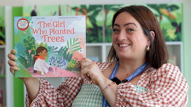 A staff member from Newington Library holding up a book called "The Girl Who Planted Trees" and smiling 