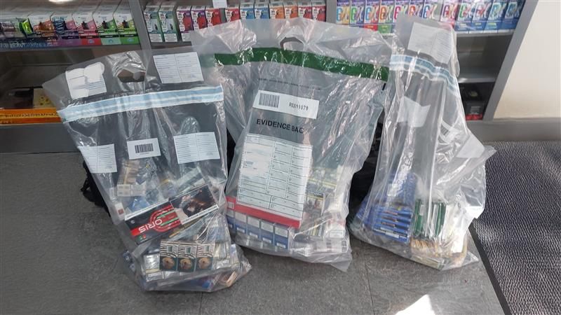 Three evidence bags full of illicit cigarettes sitting in front of shelves of similar products on a shop floor
