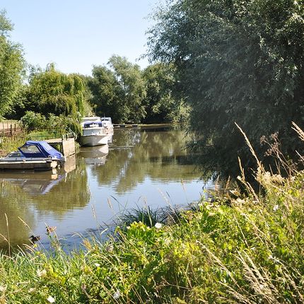 Calm river with moored boats along the bank, surrounded by lush green trees and tall grasses under a clear blue sky.