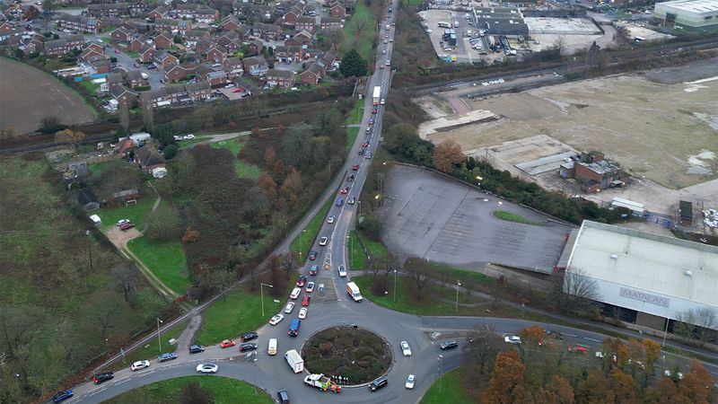 The image shows a road with a roundabout in an urban brownfield area with cars queuing