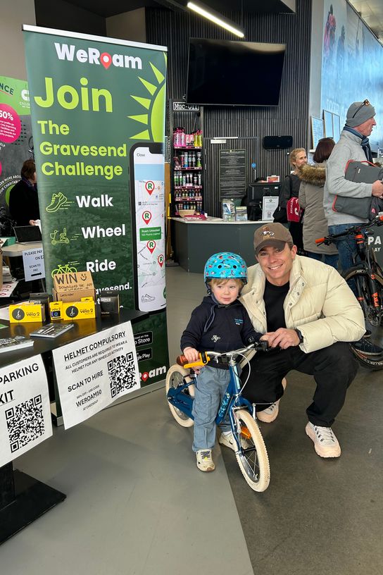 A child sat on a bike wearing a helmet and a man crouching down next to him smile towards the camera.