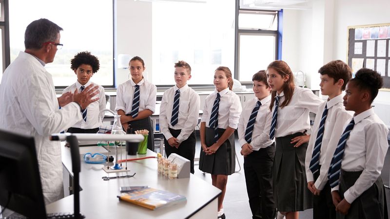 A group of secondary school children gather round a desk, listening to a teacher