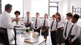 A group of secondary school children gather round a desk, listening to a teacher