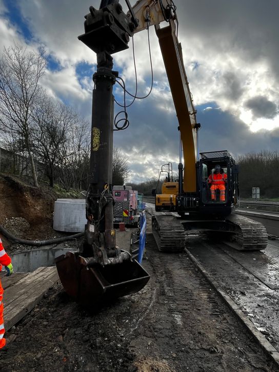 Workers in high‑visibility clothing stand near equipment and concrete drainage components, with a cloudy sky and roadside vegetation in the background.&rdquo;