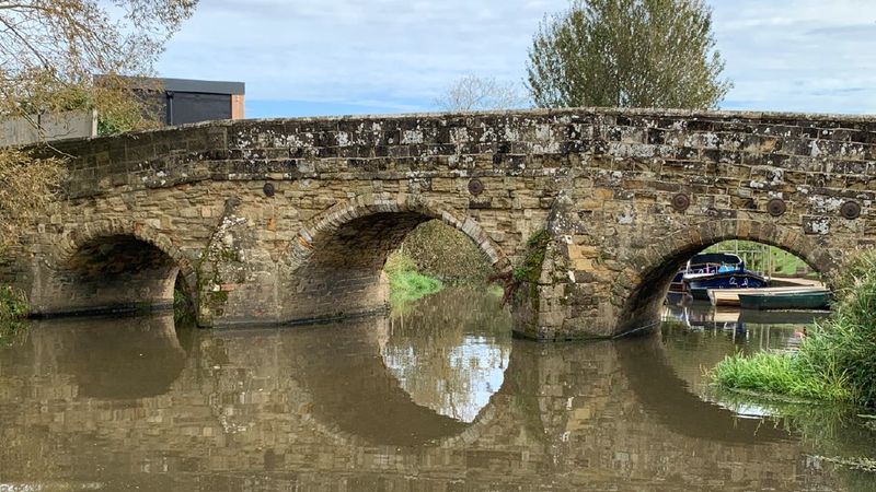 Old stone bridge with three arches spanning a calm canal, with reflections in the water and moored boats visible through the right arch.
