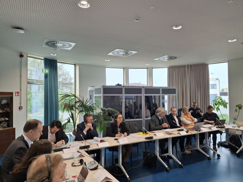 A formal meeting taking place in a modern conference room, with several people seated around joined tables. Participants are using microphones, headsets, papers and laptop computers. Large windows line the room, with plants and a glass interpretation booth visible in the background.