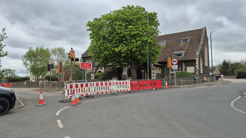 A road junction closed with red-and-white barriers and cones, traffic lights switched off, and a large leafy tree and stone building behind the closure.
