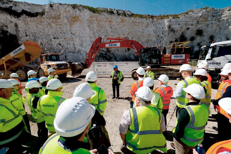 A group of people wearing high‑visibility jackets and white safety helmets stand on a construction site, listening to a briefing in front of excavators and construction vehicles, with a chalk cliff face in the background.