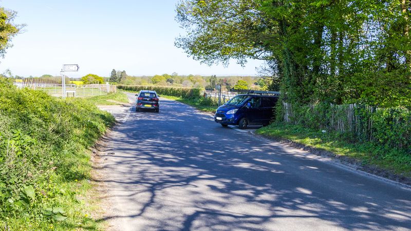 A photograph of a road with a car driving along it and at the junction a van is trying to come out.