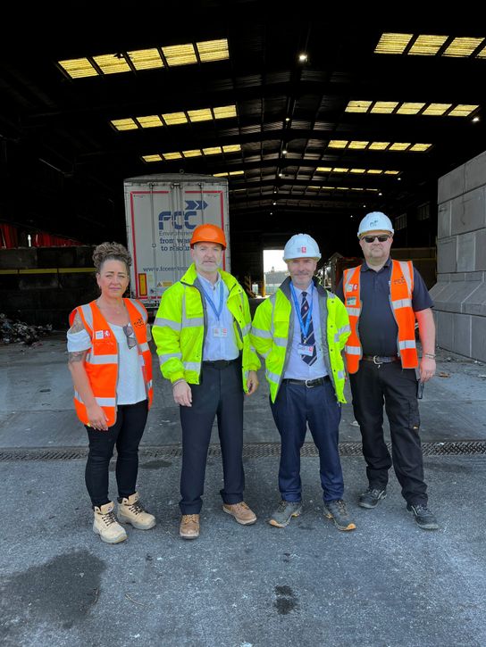 Four people wearing high-visibility clothing and hard hats stand side by side inside a large industrial warehouse. Behind them is a waste truck with &ldquo;FCC Environment &ndash; from waste to resource&rdquo; branding, along with piles of materials and heavy equipment. The building has a high metal roof with skylights letting in daylight, and the concrete floor shows scattered debris typical of a recycling or waste-processing facility.