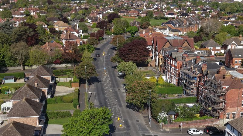 Aerial view of Shorncliffe Road, a residential neighbourhood, with tree-lined streets and detached houses, centred on a multi-lane road junction with traffic islands and pedestrian crossings. Brick houses with pitched roofs and front gardens line both sides of the road, with several parked and moving cars visible. On the right, a large red-brick building is partly covered in scaffolding, while the background shows a dense spread of similar homes and green spaces.