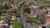 Aerial view of Shorncliffe Road, a residential neighbourhood, with tree-lined streets and detached houses, centred on a multi-lane road junction with traffic islands and pedestrian crossings. Brick houses with pitched roofs and front gardens line both sides of the road, with several parked and moving cars visible. On the right, a large red-brick building is partly covered in scaffolding, while the background shows a dense spread of similar homes and green spaces.